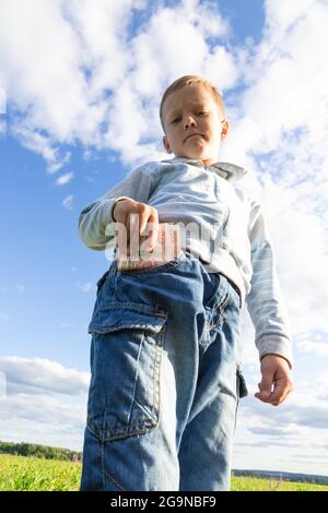 Un enfant d'âge préscolaire satisfait en jeans et un chandail tient l'argent de papier dans la nature dans un champ sur le fond d'un ciel bleu avec des nuages. Portrait Banque D'Images