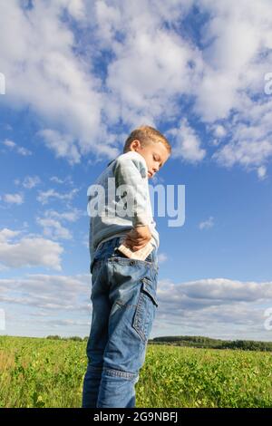 Un enfant d'âge préscolaire satisfait en jeans et un chandail tient l'argent de papier dans la nature dans un champ sur le fond d'un ciel bleu avec des nuages. Portrait Banque D'Images