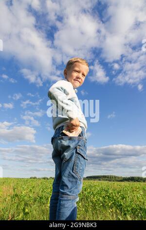 Un enfant d'âge préscolaire satisfait en jeans et un chandail tient l'argent de papier dans la nature dans un champ sur le fond d'un ciel bleu avec des nuages. Portrait Banque D'Images