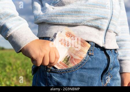 La main d'un enfant prend de l'argent en papier roubles d'une poche de Jean sur un fond de ciel bleu lors d'une journée ensoleillée d'été. Mise au point sélective. Gros plan Banque D'Images