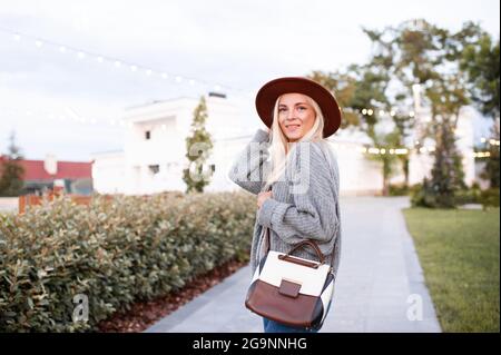 Sourire belle blonde adolescente 17-18 ans porter chapeau élégant, sac en cuir et pull gris tricoté marcher dans la rue de la ville dans le parc à l'extérieur. Réglage Banque D'Images