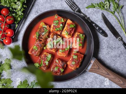 À plat, déposer les petits pains de chou farcis, la sauce tomate et le persil haché sur la poêle. Tomates, persil, fourchette et couteau sur le côté de la pierre texturée grise Banque D'Images