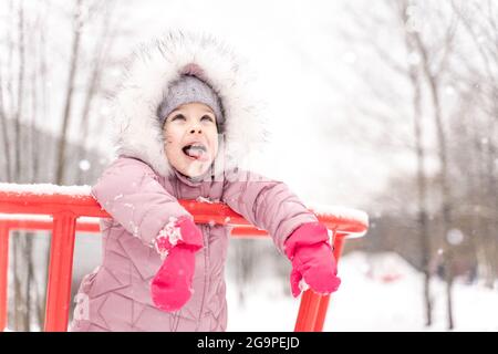 Petite fille drôle en rose vêtements d'hiver marche pendant une chute de neige. Activités d'hiver en plein air pour les enfants. Enfant mignon attrapant des flocons de neige avec sa langue Banque D'Images