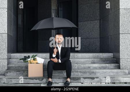 Concept de chômage. Homme d'affaires mature sans emploi assis sous un parapluie avec une affiche vide et une boîte d'effets personnels Banque D'Images