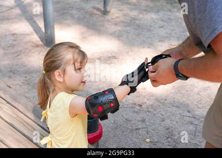 Père papa mettant des protège-coudes et des genouillères pour le vélo sur sa fille. Aider et prendre soin des enfants. Sécurité urbaine sur scooter ou vélo Banque D'Images