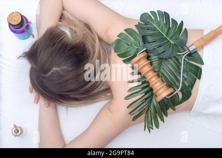 Madero thérapie, massage relaxant - masseur à roulettes reposant sur des feuilles vertes sur le dos d'une femme et des bouteilles avec des huiles essentielles, gros plan, vue du dessus. Corps c Banque D'Images