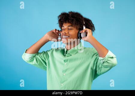 Chanson préférée. Un homme afro-américain décontracté dans un casque qui écoute de la musique avec les yeux fermés, debout sur fond bleu Banque D'Images