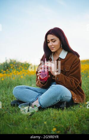 Une adolescente boit de l'eau dans un pot rouge assis sur l'herbe d'une colline. Elle a une salade à côté d'elle. Banque D'Images