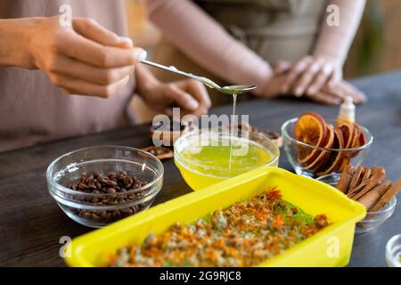 Mains de la femme versant la masse de savon fondu dans un contenant en silicone avec des ingrédients naturels secs tout en faisant des produits cosmétiques à la maison Banque D'Images