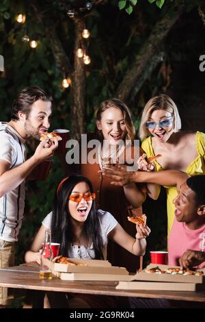 Homme afro-américain qui pousse ses amis loin de la pizza près de la bière sur la table à l'extérieur Banque D'Images