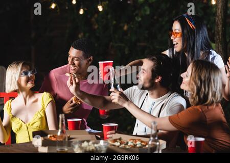 Des amis interraciaux souriants tenant une pizza près d'une femme et des tasses en plastique sur une table à l'extérieur Banque D'Images