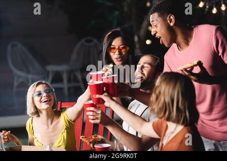 Des gens interraciaux excités qui toaster avec des tasses en plastique et qui tiennent des pizzas à l'extérieur le soir Banque D'Images