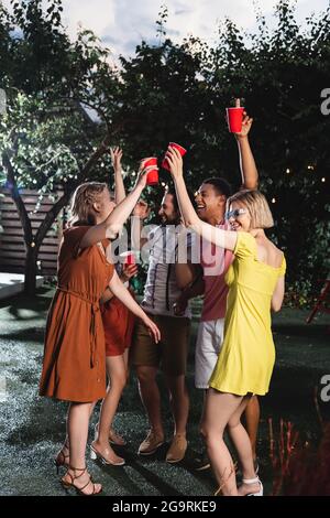 Des personnes multiethniques enthousiastes élèvent des tasses en plastique pendant la fête le soir Banque D'Images