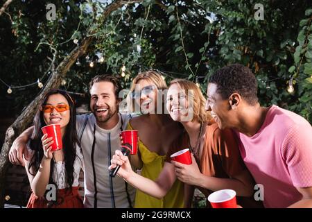 des amis multiethniques heureux avec des tasses en plastique chantant karaoké dans le jardin de nuit Banque D'Images