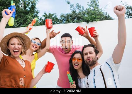 des amis multiethniques enthousiastes avec des tasses en plastique et des pistolets à eau montrant du rock et gagnez des gestes pendant la fête Banque D'Images