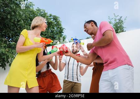 joyeux amis multiculturels en dégustant des tasses en plastique tout en tenant des pistolets à eau Banque D'Images