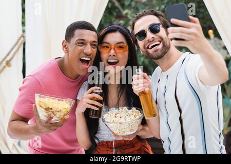 des amis multiethniques enthousiastes qui prennent du selfie avec de la bière et des collations sur leur téléphone portable Banque D'Images