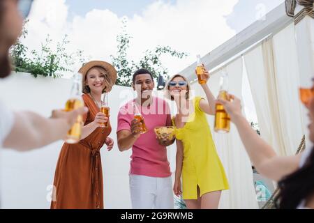 des amis multiculturels excités qui se réjouissent de déguster des bouteilles de bière pendant la fête sur un premier plan flou Banque D'Images