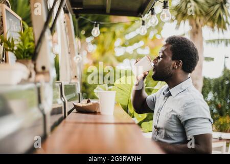 Jeune africain mangeant et buvant dans un comptoir de camion de nourriture en plein air dans le parc de la ville - Focus sur le visage. Banque D'Images