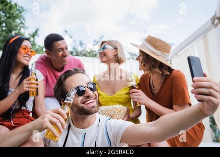 homme joyeux prenant le selfie sur smartphone pendant la fête estivale avec des amis multiethniques flous Banque D'Images