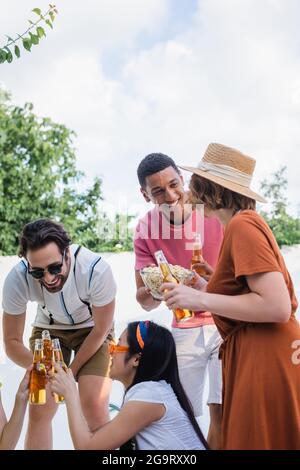 un homme afro-américain enthousiaste tenant du pop-corn près d'amis multiethniques en dégustant des bouteilles de bière Banque D'Images