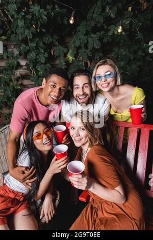 Des amis interraciaux souriants avec des tasses en plastique qui s'embrasent et regardent l'appareil photo à l'extérieur Banque D'Images