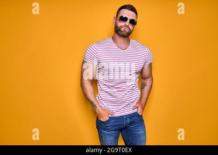 Un homme à barbe musclé et confiante en chemise, Jean et lunettes de soleil stylées debout en studio sur fond jaune Banque D'Images