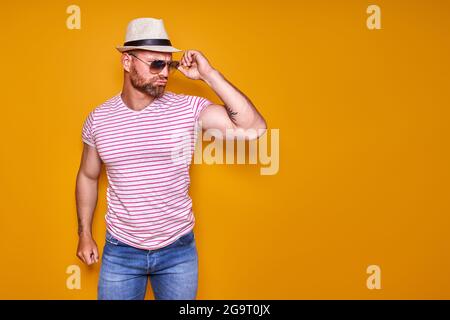 Homme musclé à barbe en chemise, Jean et chapeau élégant, lunettes de soleil debout en studio sur fond jaune Banque D'Images