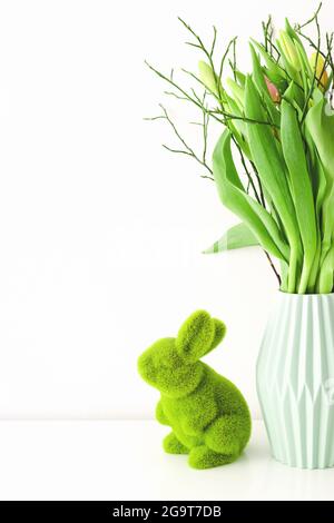 Bouquet de tulipes multicolores non ouvertes dans un vase en céramique vert clair et lapin en herbe vert de Pâques sur fond blanc. Concept de décoration de vacances. Banque D'Images