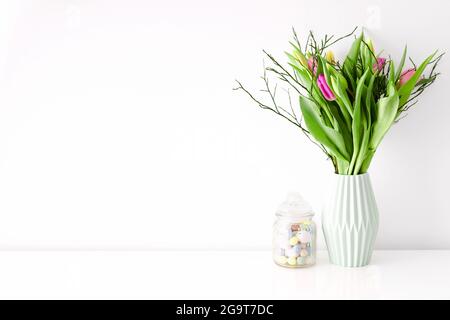 Bouquet de tulipes multicolores non ouvertes dans un vase en céramique vert clair et un pot de friandises de Pâques rempli d'œufs sur fond de mur blanc. Décoration intérieure Banque D'Images