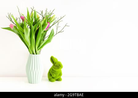 Bouquet de tulipes multicolores non ouvertes dans un vase en céramique vert clair et lapin en herbe vert Pâques sur fond de mur blanc. Concept de décoration de vacances. Banque D'Images