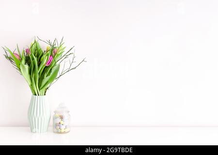 Bouquet de tulipes multicolores non ouvertes dans un vase en céramique vert clair et un pot de friandises de Pâques rempli d'œufs sur fond de mur blanc. Décoration intérieure Banque D'Images