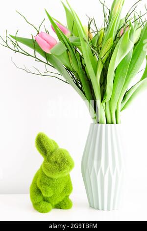 Bouquet de tulipes multicolores non ouvertes dans un vase en céramique vert clair et lapin en herbe vert Pâques sur fond de mur blanc. Concept de décoration de vacances. Banque D'Images