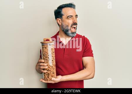 Homme d'âge moyen avec la barbe et les cheveux gris tenant le pot avec des pâtes macaroni en colère et fou criant frustré et furieux, criant avec la colère. Rage et Banque D'Images