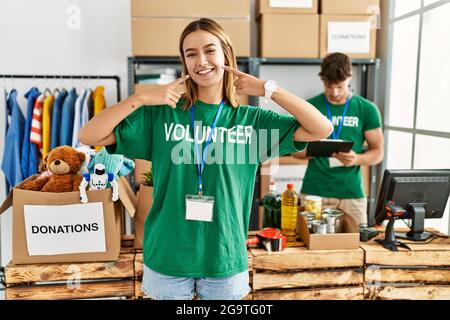 Jeune fille blonde portant le t-shirt de bénévole au stand de don souriant gai montrant et pointant avec les doigts dents et la bouche Banque D'Images