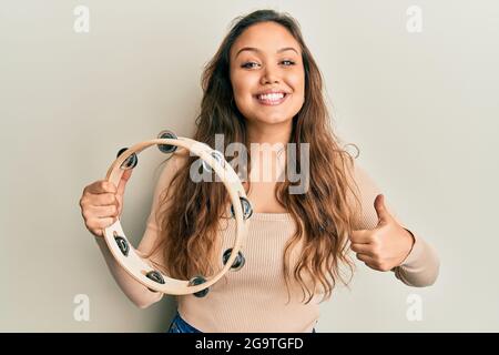 Jeune fille hispanique jouant tambourine souriant heureux et positif, pouce vers le haut faisant excellent et signe d'approbation Banque D'Images