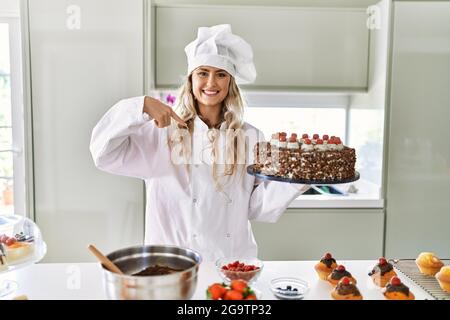 Jeune chef de pâtisserie caucasienne femme cuisant des pâtisseries et des gâteaux à la cuisine pointant du doigt à un soi souriant heureux et fier Banque D'Images