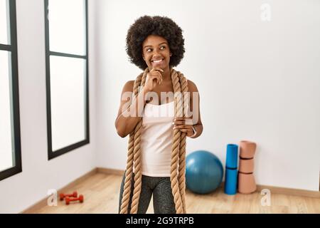 Jeune afro-américaine avec des cheveux afro à la salle de gym avec des cordes de bataille avec la main sur le menton en pensant à la question, l'expression pensive. Smili Banque D'Images