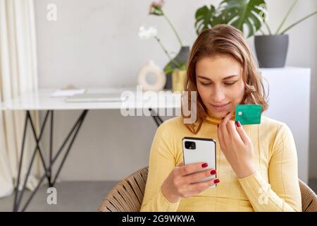 Elle a été ravie d'acheter une carte en plastique et un smartphone chez elle. Femme souriante payant par carte pour un achat en ligne par téléphone Banque D'Images