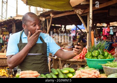 Photo de stock de l'épicerie africaine masculine avec un tablier, étonné comme il se tient et fixe à son téléphone Banque D'Images