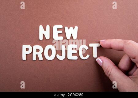 NOUVEAU PROJET. Lettres blanches en bois avec texte et main de femme sur fond marron. Banque D'Images