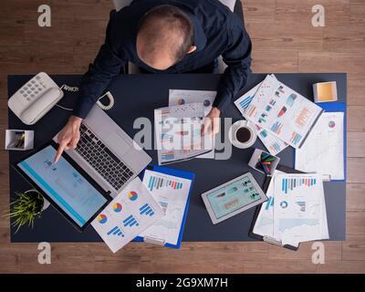 Vue de dessus de l'homme d'affaires pointant sur les statistiques financières sur l'écran d'ordinateur portable assis au bureau d'entreprise avec paperwork. Employé utilisant une tablette pc avec des graphiques. Banque D'Images