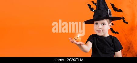 Un garçon en costume de sorcière dans un chapeau noir tient une petite lampe de poche à citrouille Jack. Sur un fond orange. Halloween enfants. Emplacement de la bannière pour Banque D'Images