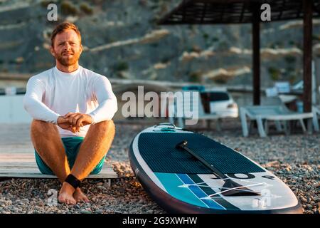 Beau homme bronzé assis à la plage de galets avec un panneau de soutien. Parapluies en bois et chaise longue sur fond défoqué. Surf et activité. Banque D'Images