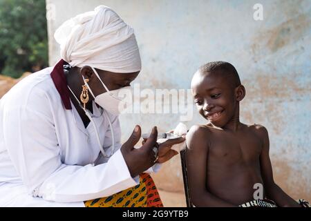 Dans cette image, une infirmière noire avec masque est en train d'injecter une dose de vaccin à un garçon d'école courageux souriant dans un milieu hospitalier africain Banque D'Images