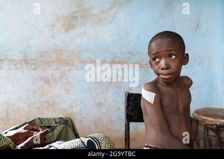 Dans cette image, un petit garçon noir malheureux d'Afrique assis sur une chaise avec un grand plâtre sur son bras où il vient de recevoir un vaccin douloureux est l Banque D'Images