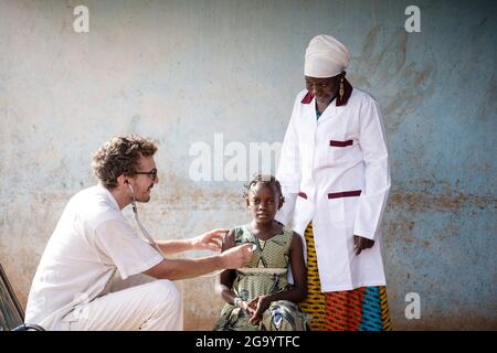 Dans cette image, un médecin blanc rassurant auscultation la poitrine d'une petite fille africaine sérieuse avec une infirmière noire qui le regarde avec joie Banque D'Images