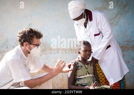 Dans cette image, un médecin pédiatre caucasien vaccine une petite fille africaine courageuse soutenue par une infirmière noire aimable qui tient ses sholders. Banque D'Images