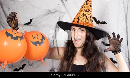Une femme en costume de sorcière dans un chapeau et des gants à l'arrière-plan avec des chauves-souris tient des balles de citrouille et dit boo. Bannière longue pour Halloween. Banque D'Images