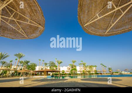 Vue sur les parasols en paille contre le ciel bleu avec vue sur la piscine de l'hôtel et les palmiers en Egypte Banque D'Images